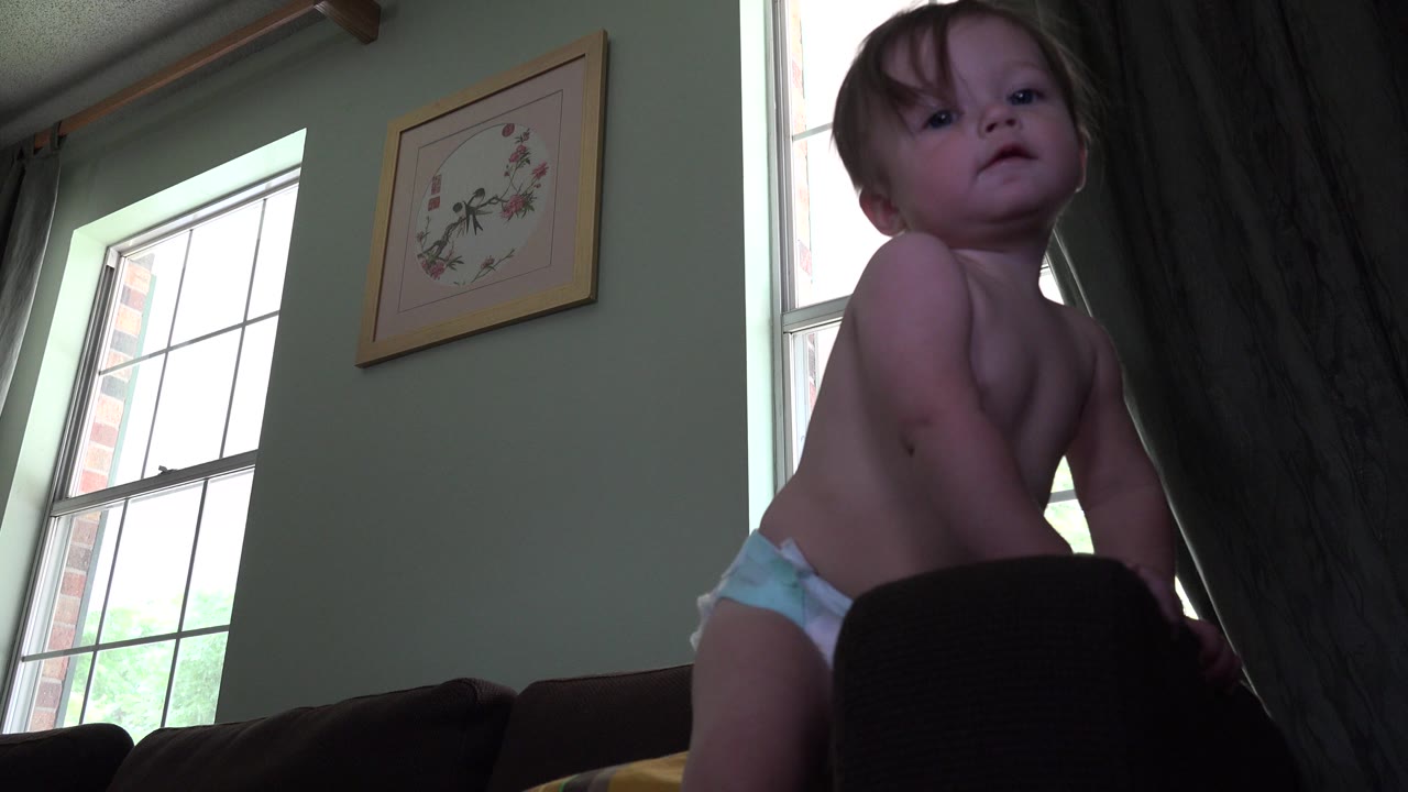 A close low-angle view shows the blond baby in Westminster, Colorado with hair partly across the forehead, dark furniture in front, and tall windows in the background.