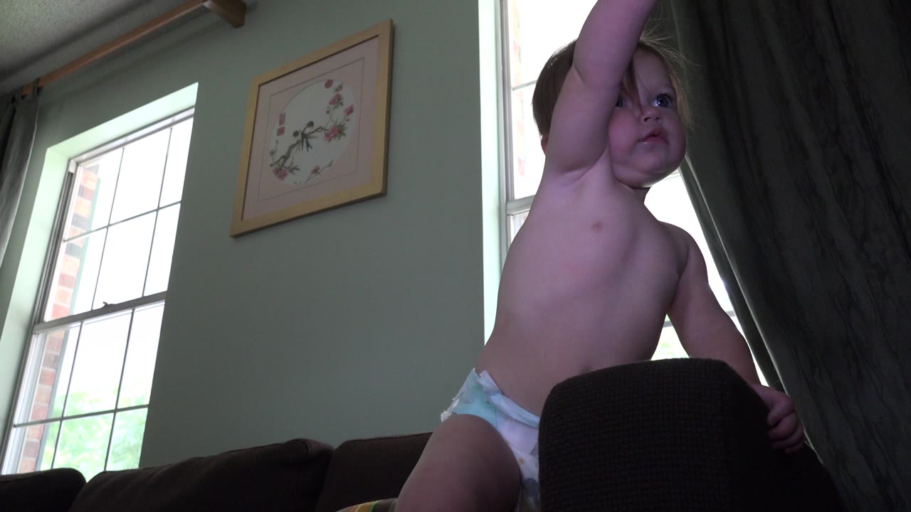 Toddler in a diaper standing on a couch near tall windows.