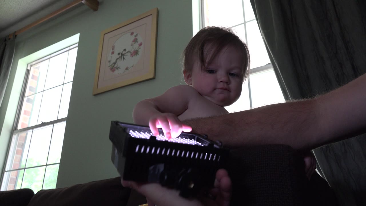 Baby reaches toward a glowing LED panel beside an adult arm.