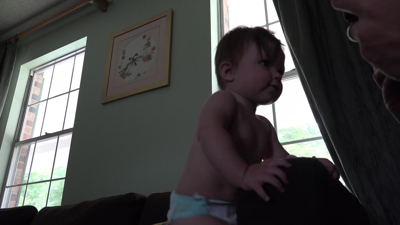 Toddler standing near a couch by a bright window as an adult face appears at the edge.