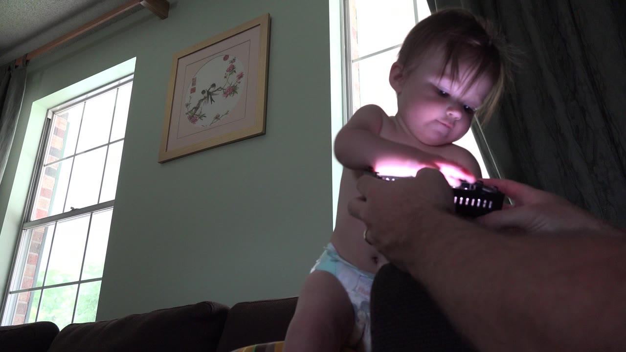 The toddler looks down toward a dark handheld object held by adult hands, with bright window light outlining the living room in Westminster, Colorado.
