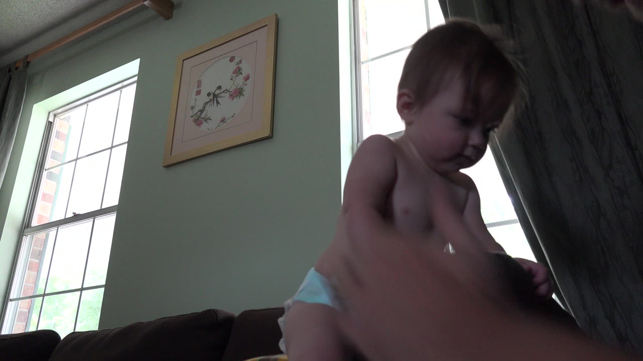 Baby girl standing near an adult hand in a living room.