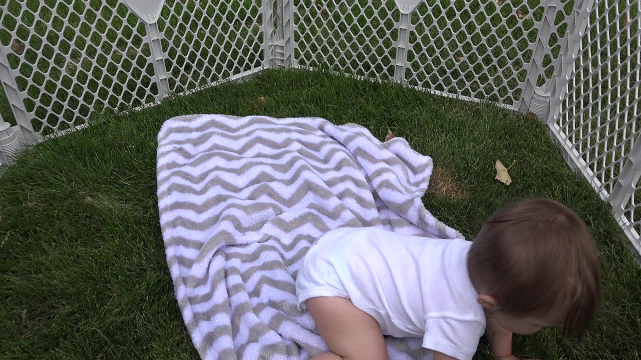 Baby crawling beside a chevron blanket on grass inside a white safety fence.