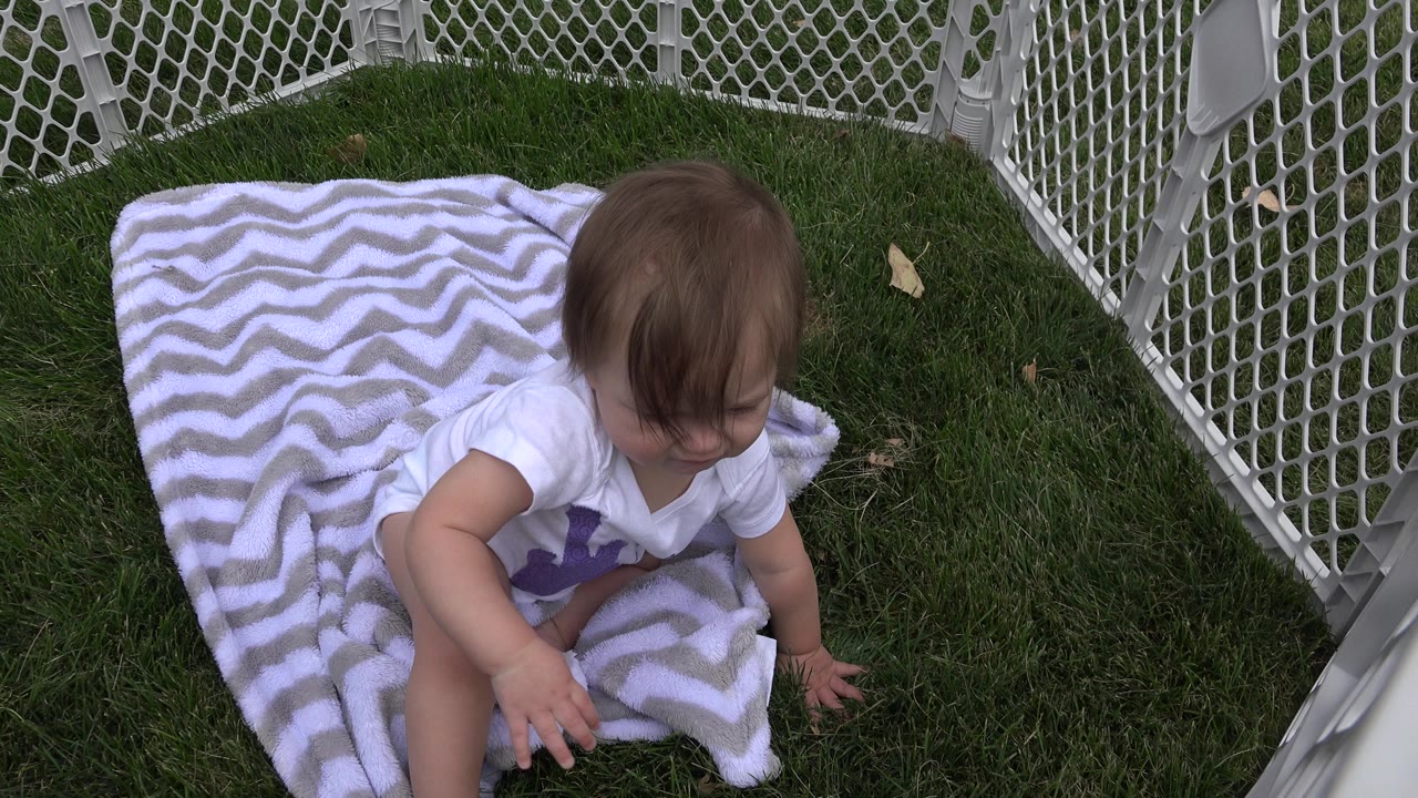 Baby seated on a striped blanket inside a white playpen on grass.