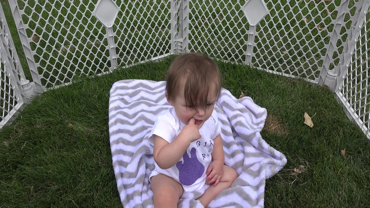 Young child sitting on a striped blanket inside a white baby fence on grass.
