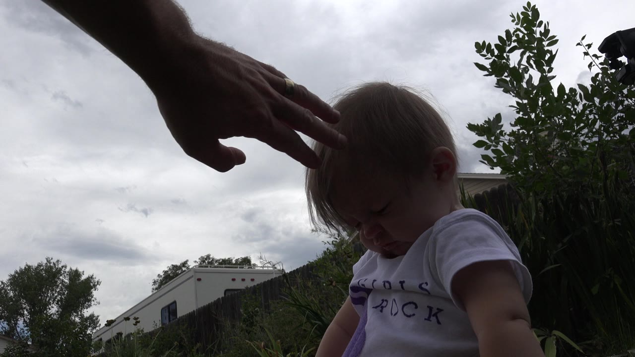 Adult hand above a baby in a white shirt outdoors.