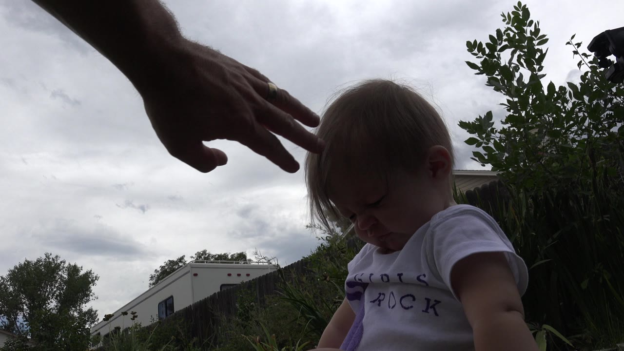 In Westminster, Colorado, the baby remains low in frame as an adult hand passes above the child's head, with gray sky and yard greenery in the background.