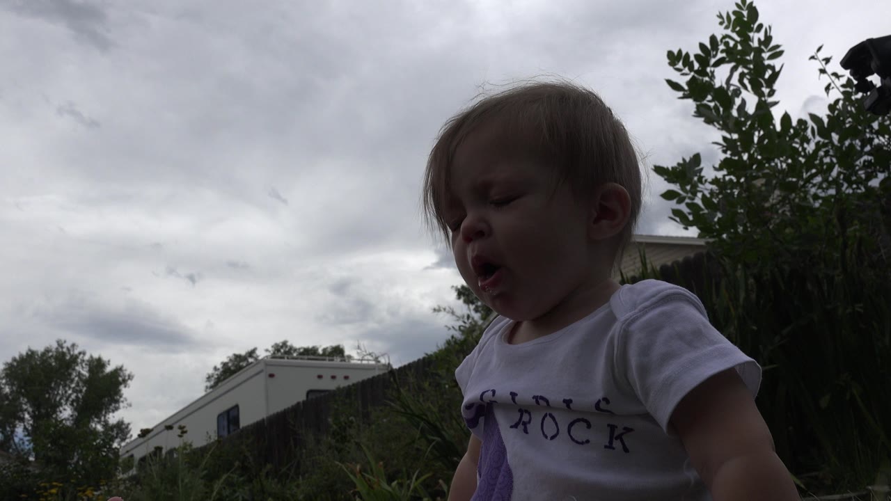 At 0:00 in Westminster, Colorado, a baby in a white T-shirt leans near a low camera with cloudy sky, trees, and a pale structure behind.