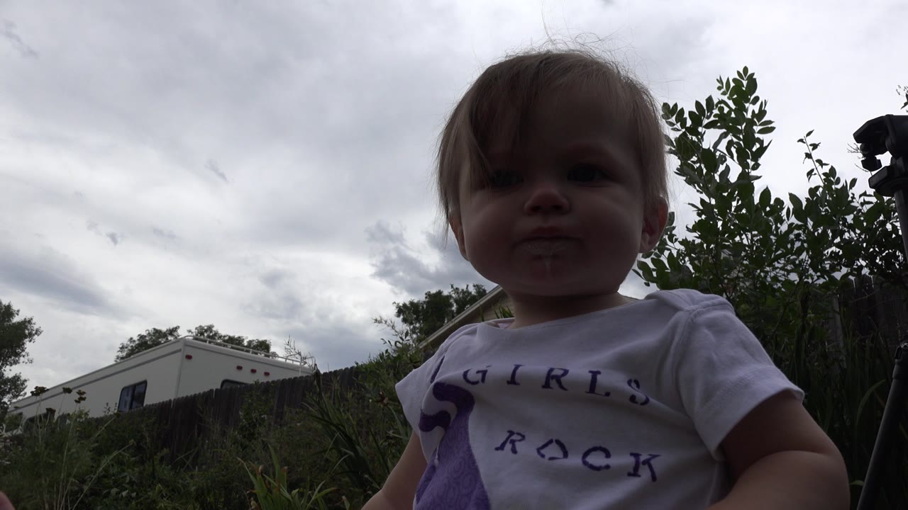 The child remains centered near the lens in Westminster, Colorado, with her face shadowed by backlight and a white building visible beyond the yard.