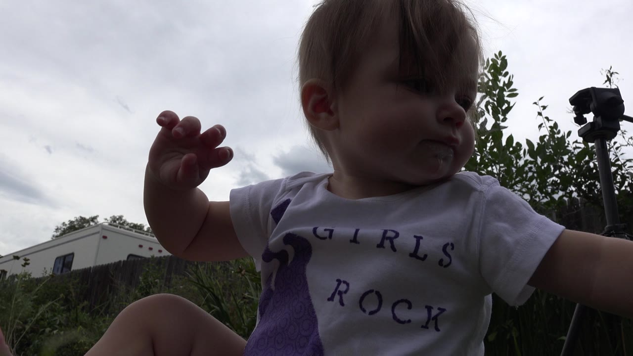 The toddler remains close to the ground in the Westminster yard, one hand lifted near her face while the fence, shrubs, cloudy sky, and a tripod or camera stand in the background.