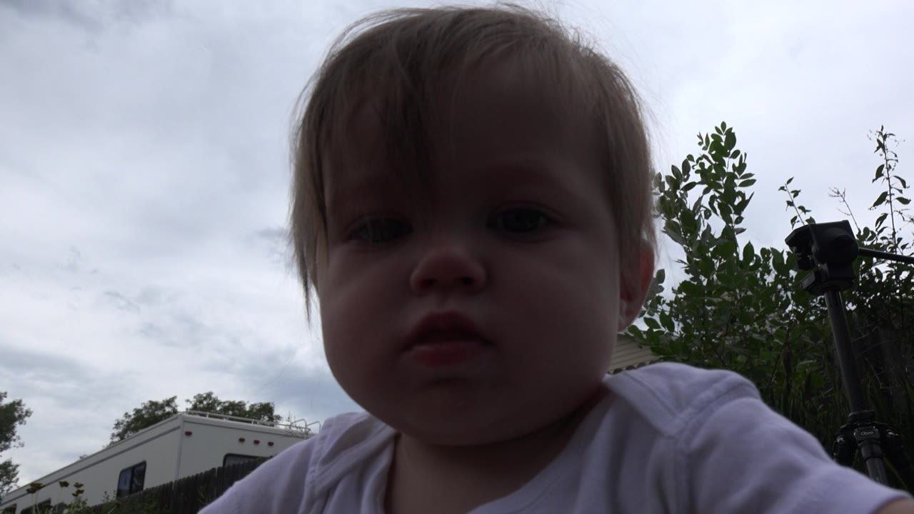 Baby girl close to a low outdoor camera under cloudy sky.