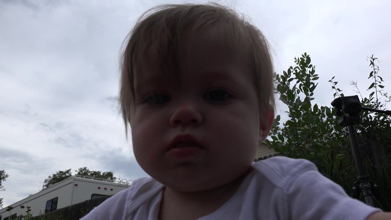The baby girl remains centered near the lens in Westminster, Colorado, with her face in shadow against a bright cloudy sky and a tripod visible over her shoulder.