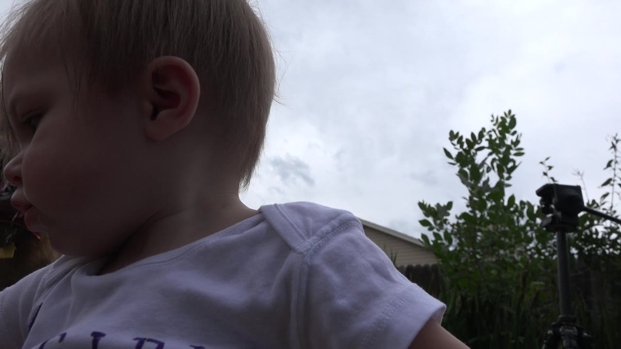 A close, low-angle view of a baby in a white shirt at the left edge of the frame in Westminster, Colorado, with cloudy sky and backyard greenery behind.