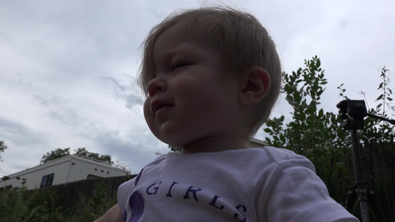 The toddler remains close to the lens in Westminster, Colorado, with a bright cloudy sky, greenery, and a dark stand visible in the background.