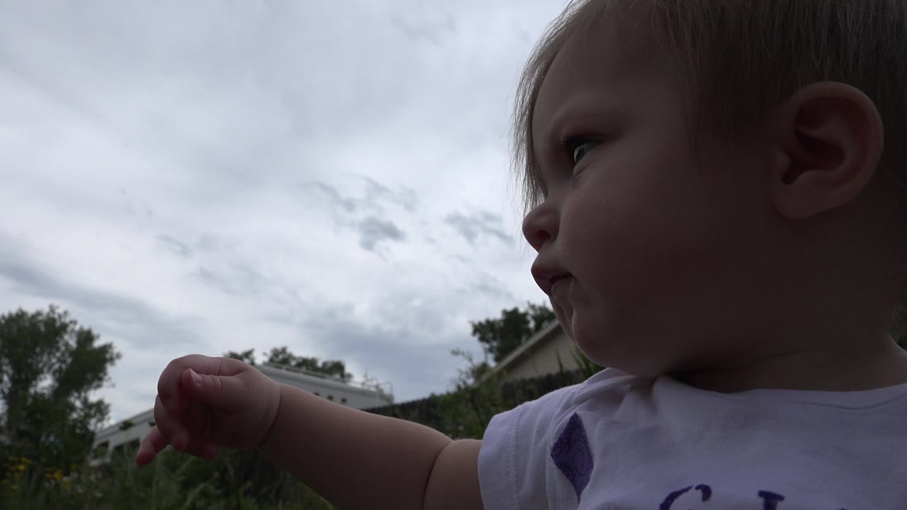 A baby in Westminster, Colorado fills the left foreground in side profile, with a cloudy sky, trees, fencing, and residential structures behind.