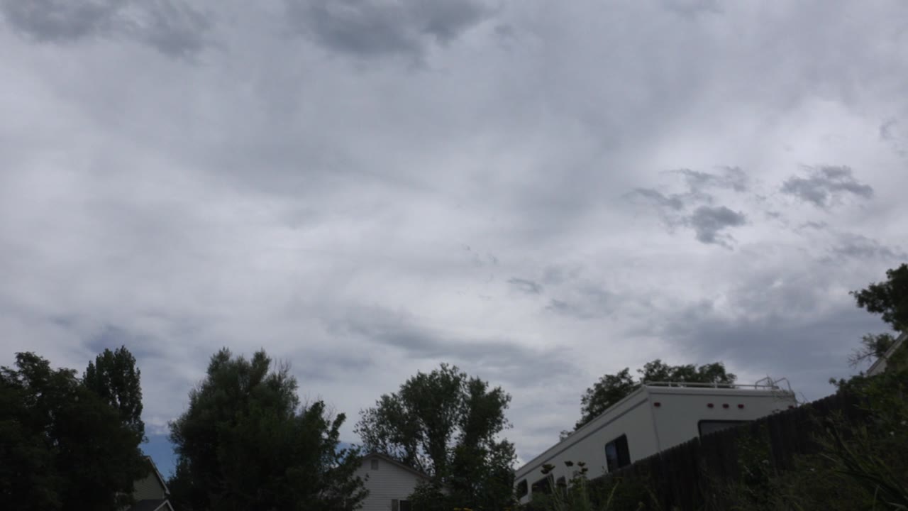 Cloudy sky over trees, fence, homes, and an RV-like vehicle.