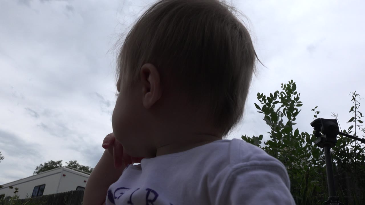 Baby in a white shirt close to the camera under a cloudy sky.