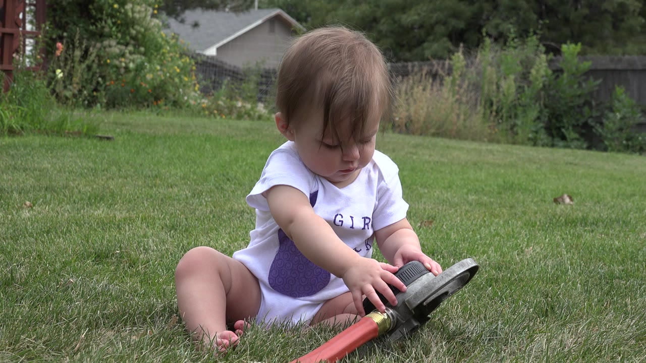 Baby sitting on grass and touching a red garden hose.