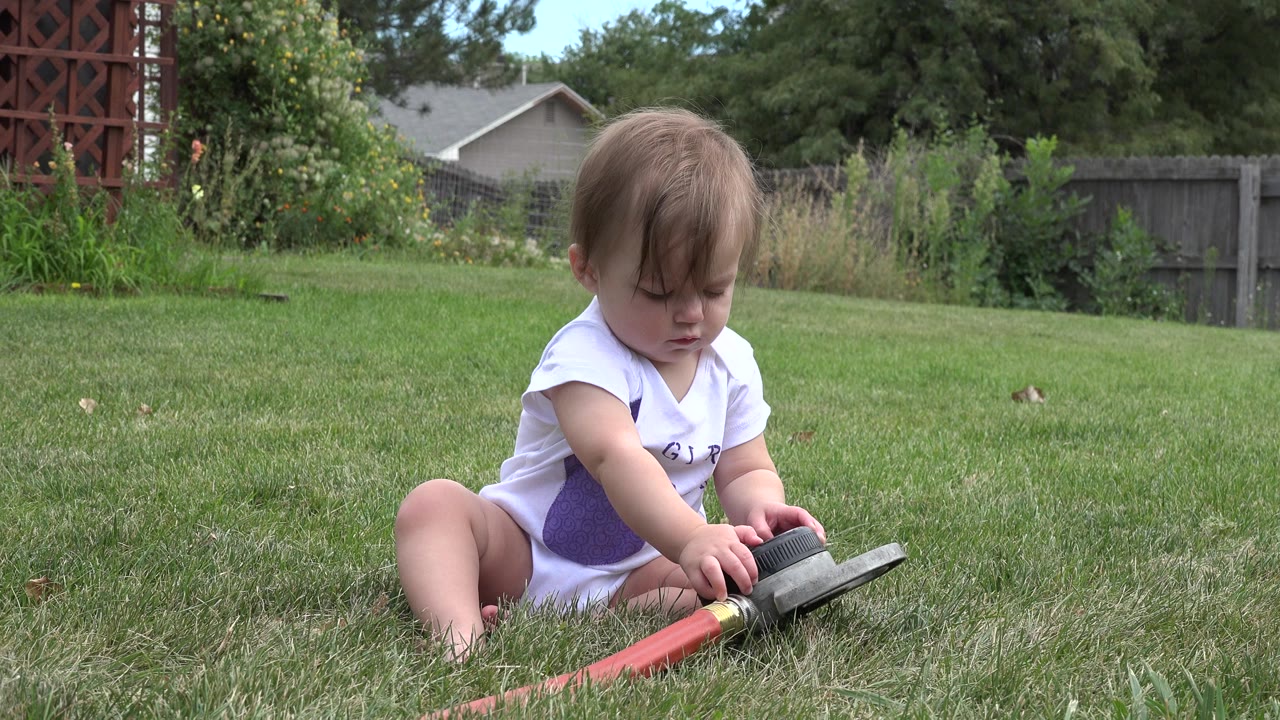 A blond baby girl sits on a Westminster, Colorado lawn, looking down while touching a red hose or springier toy in the grass.