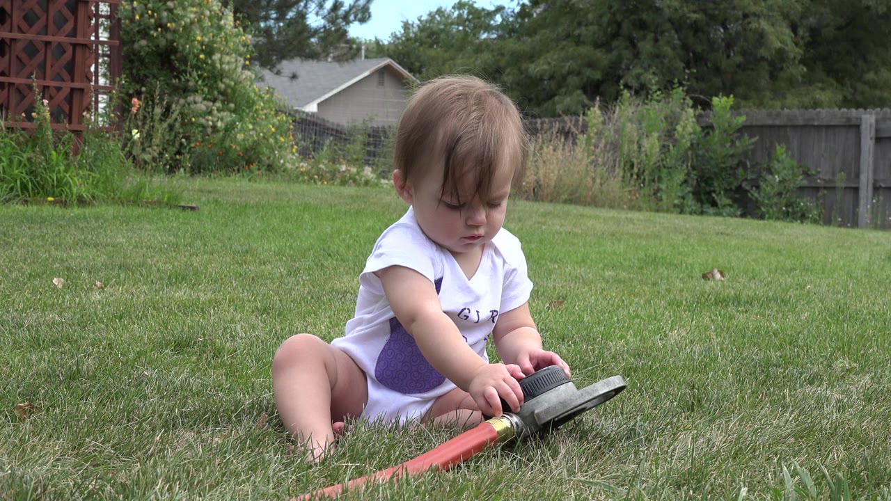 The baby girl remains seated on the grass in Westminster, Colorado, with both hands near the black end of the red hose toy.