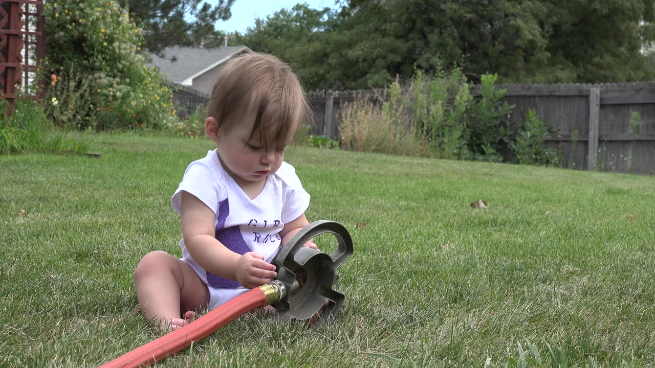 Baby girl lifting the black end of a red hose toy in the grass.