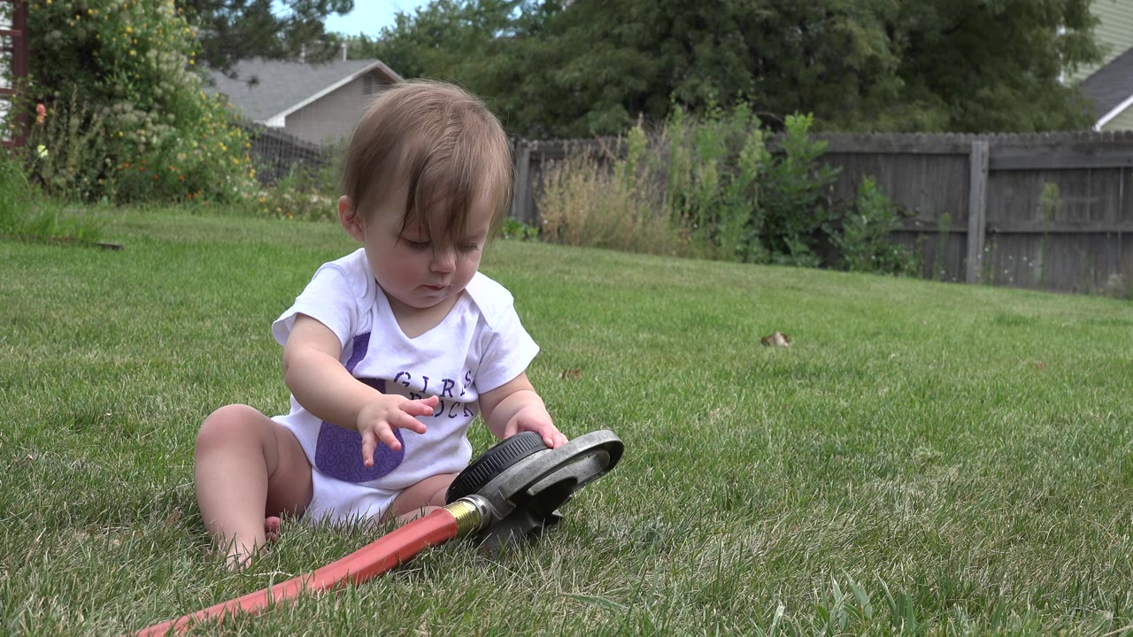 Baby girl seated on grass reaching toward a black toy attached to a red hose.
