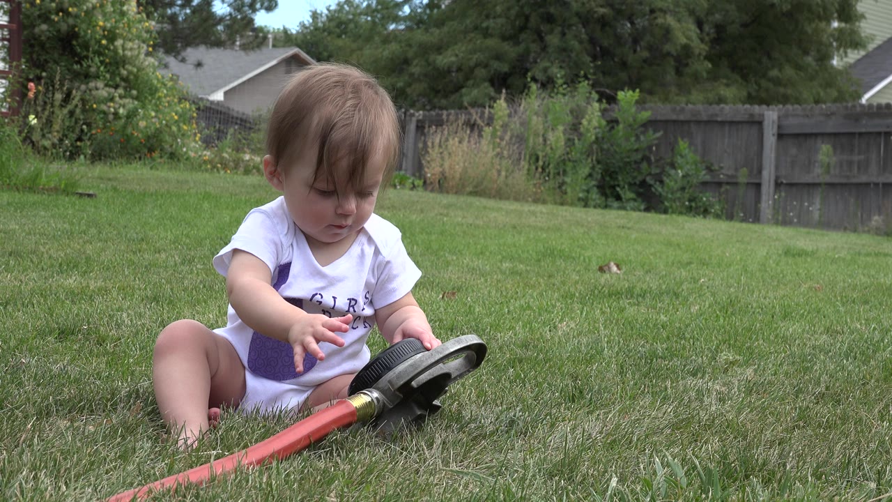 One second in, the baby girl looks down at the black yard toy while sitting on the grass in a Westminster, Colorado backyard.