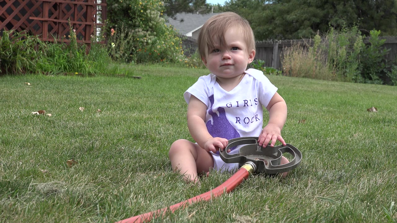 A young girl sits on a green lawn in Westminster, Colorado, facing the camera while holding a dark garden hose attachment connected to a red hose.