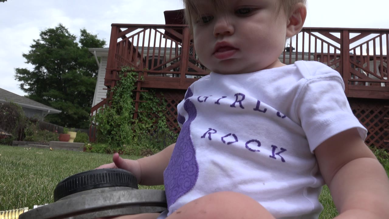Blond baby close to a ground-level camera on grass with a red deck behind.