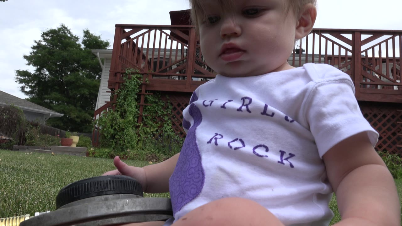 The baby remains large in the foreground in the Westminster backyard, looking downward near a dark round object at the edge of the grass while the red deck and house stay in back.