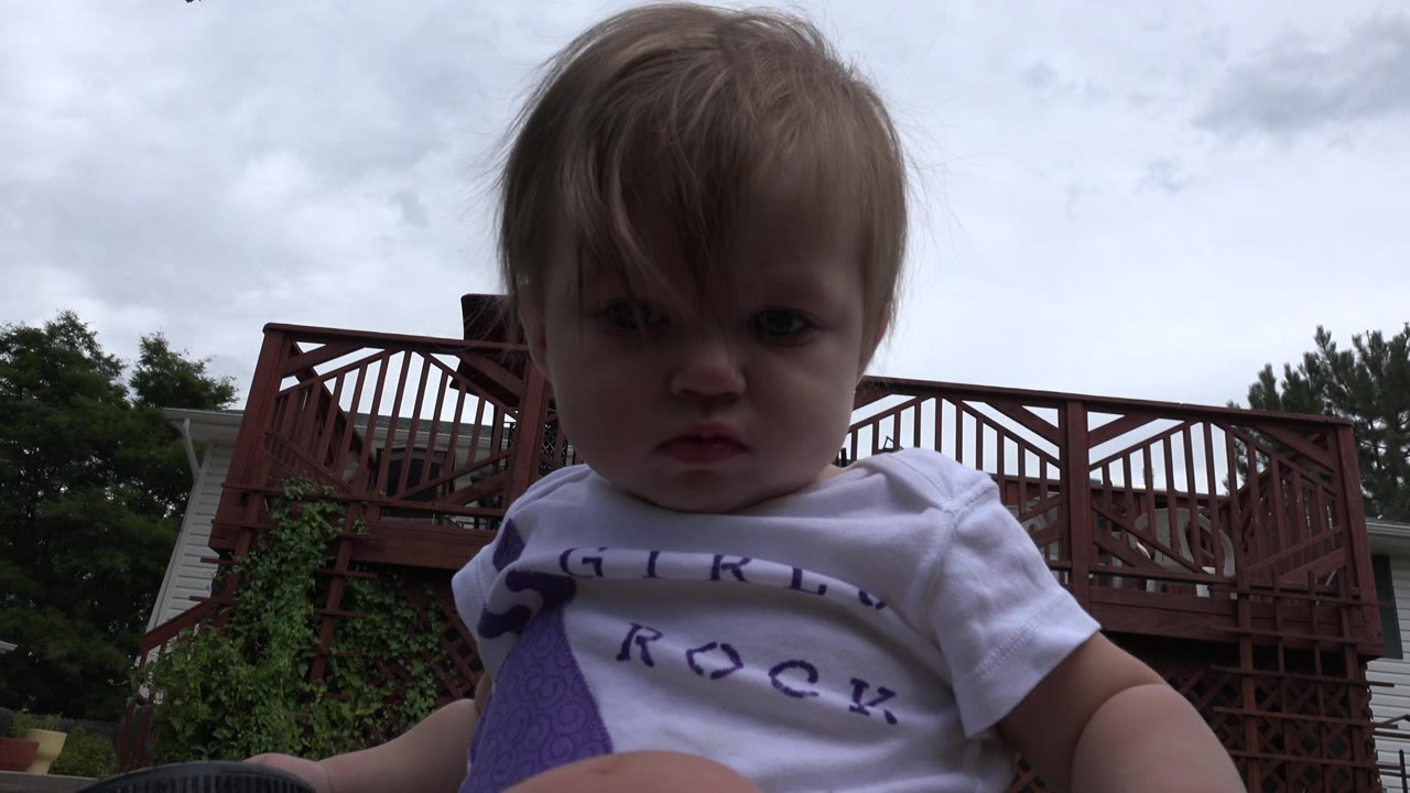 The toddler faces forward in a close low-angle frame in Westminster, Colorado, with bangs across the forehead and a wooden deck structure in the background.
