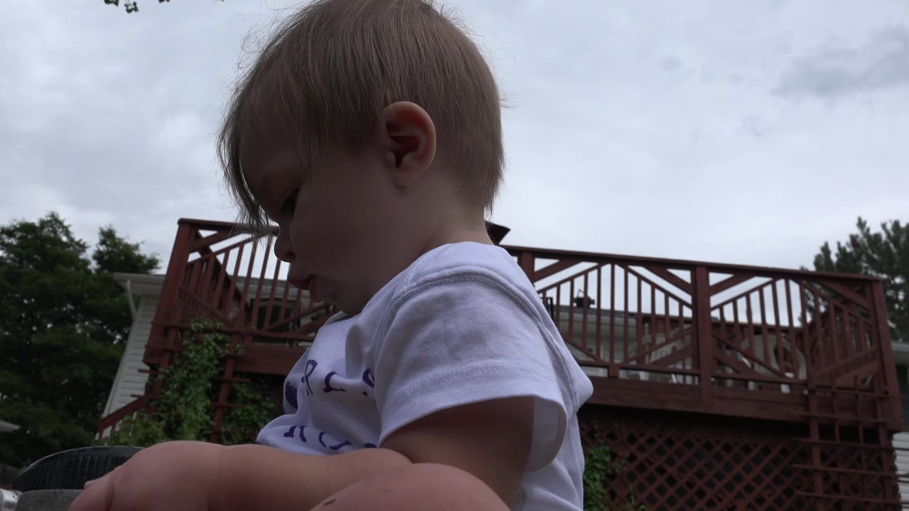 Baby in a white shirt sits low in a backyard near a red deck.