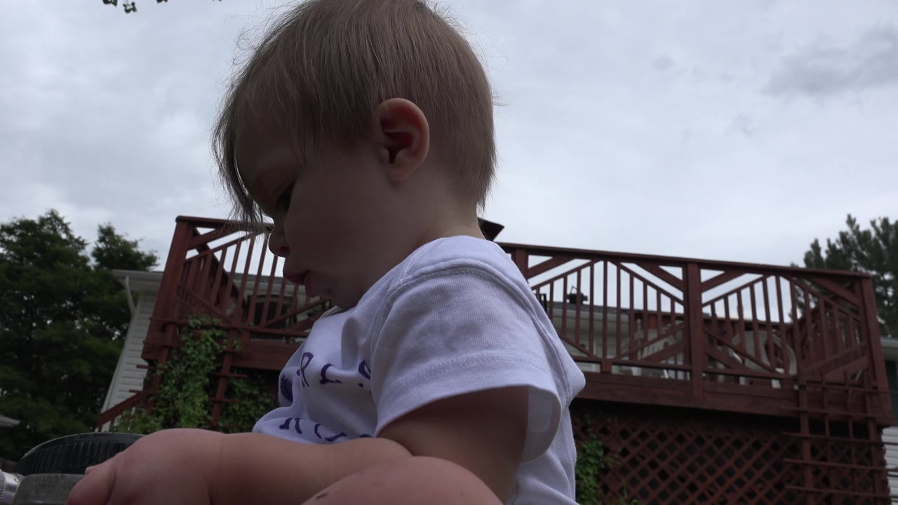 At 0:01, the baby remains in close profile looking down, with the red backyard deck and overcast sky visible across the frame.