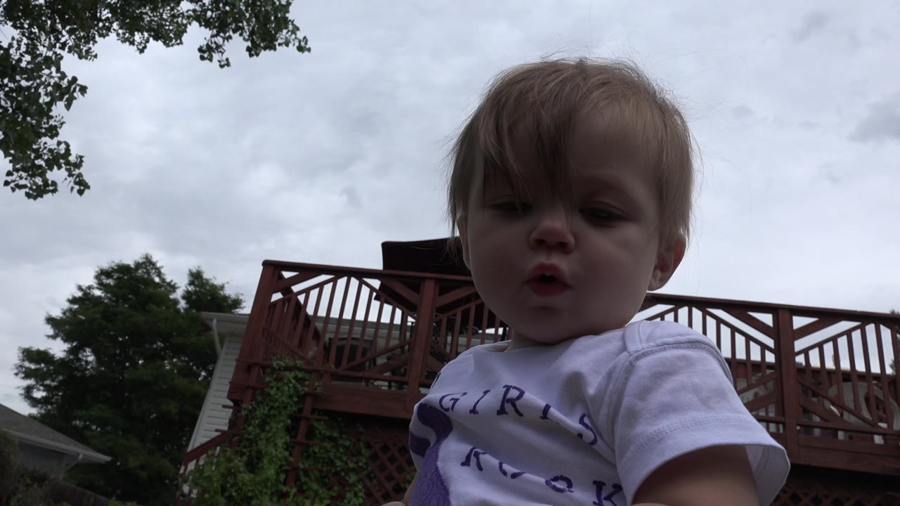 The child remains centered in a low-angle close-up, facing forward while a red wooden deck railing crosses the background in Westminster, Colorado.