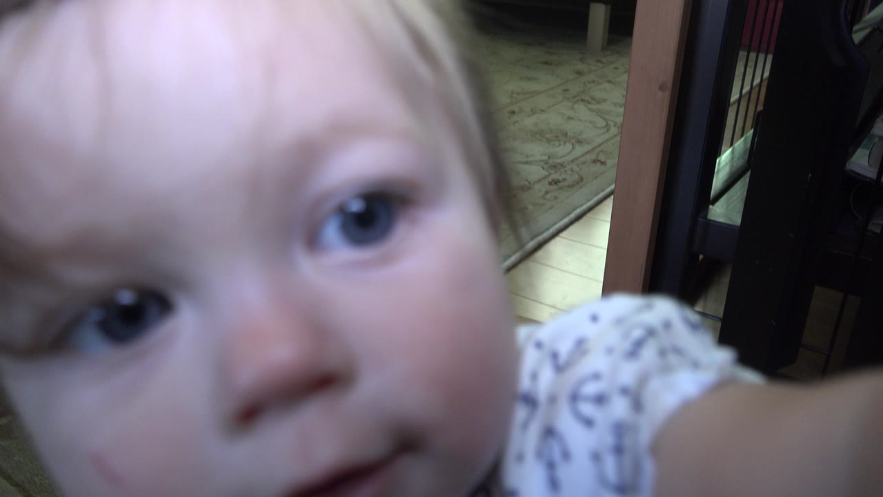 A toddler's face fills the left side of the frame indoors in Westminster, Colorado, with a patterned white shirt and dim furniture behind them.