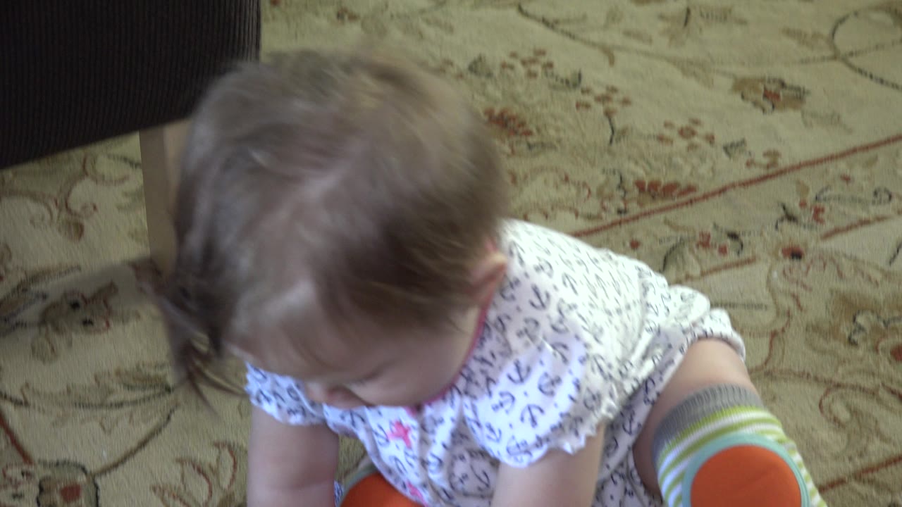 At 0:01, the baby remains seated on the patterned living room carpet, head lowered toward the hands, with gray and orange knee pads visible at the edge of the frame.