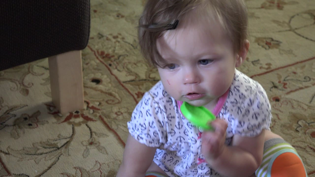 Toddler seated on a rug holding a green circular toy near her mouth.