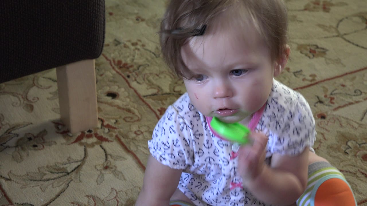 At 0:01, the toddler remains seated on the rug in Westminster, Colorado, raising the green circular toy toward her lips with furniture legs behind her.