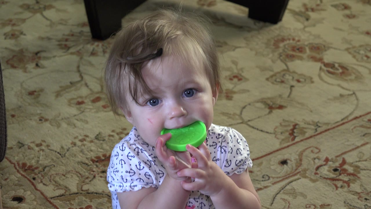 Baby holds a bright green cap near their mouth on a floral rug.