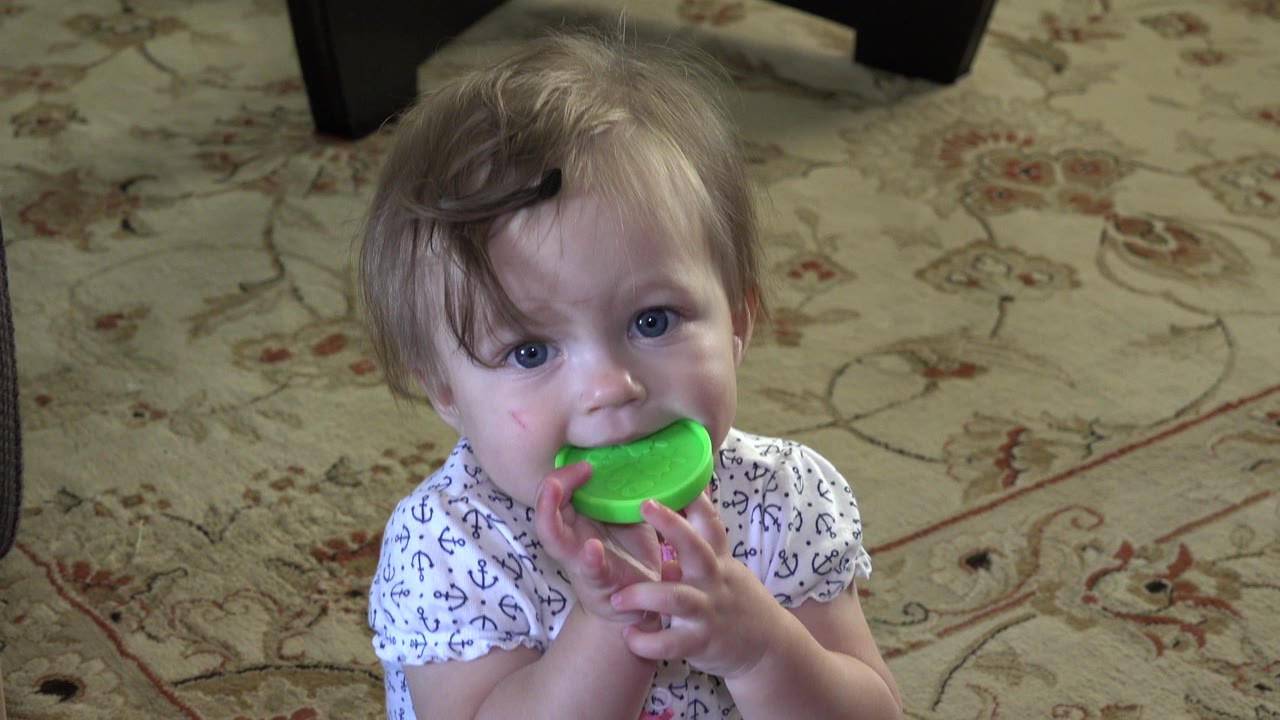 The baby remains nearly front-facing in Westminster, Colorado, with blue eyes visible and the green cap pressed against the mouth above a floral carpet.