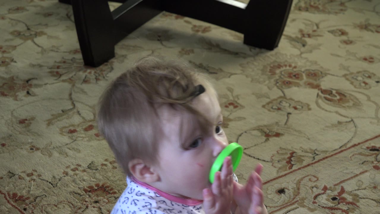 At 0:00, a blond baby in Westminster, Colorado sits near the lower left of the frame, holding a bright green toy close to the mouth on a patterned rug.