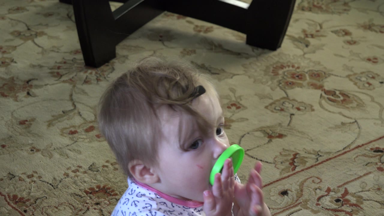 At 0:01, the baby remains seated on the patterned rug in Westminster, Colorado, with both hands around the green toy held against the mouth.
