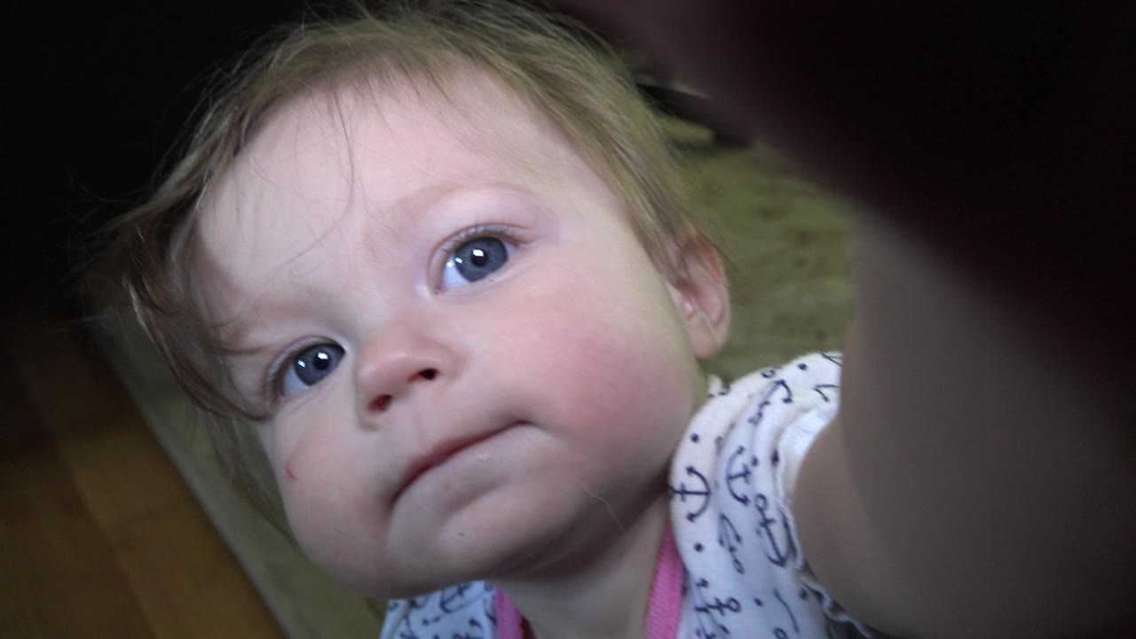 Close-up of a blonde baby girl looking up at the camera indoors.
