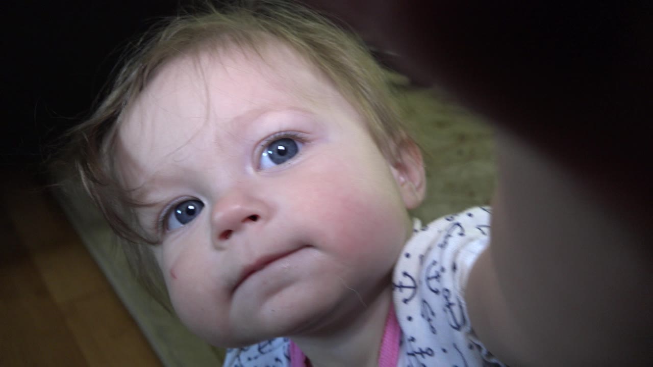 The baby girl remains close to the ground-level camera in Westminster, Colorado, with her face centered and a blurred arm or finger crossing the upper right foreground.