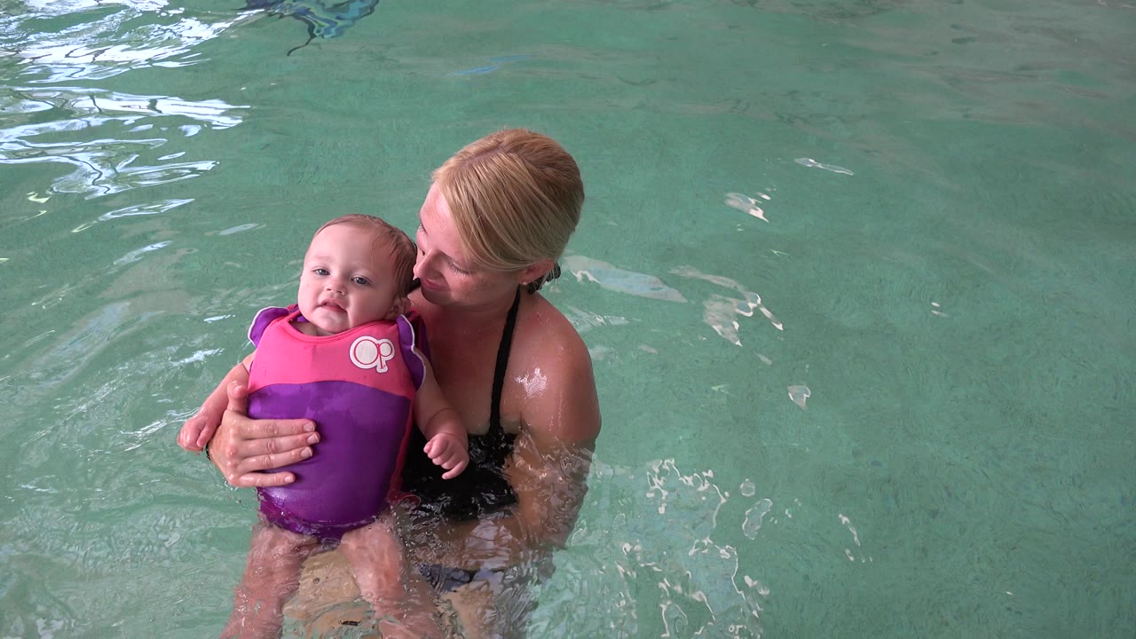 In Westminster, Colorado, an adult woman holds an infant in a pink and purple life vest near the left side of a green pool, with the infant looking toward camera.