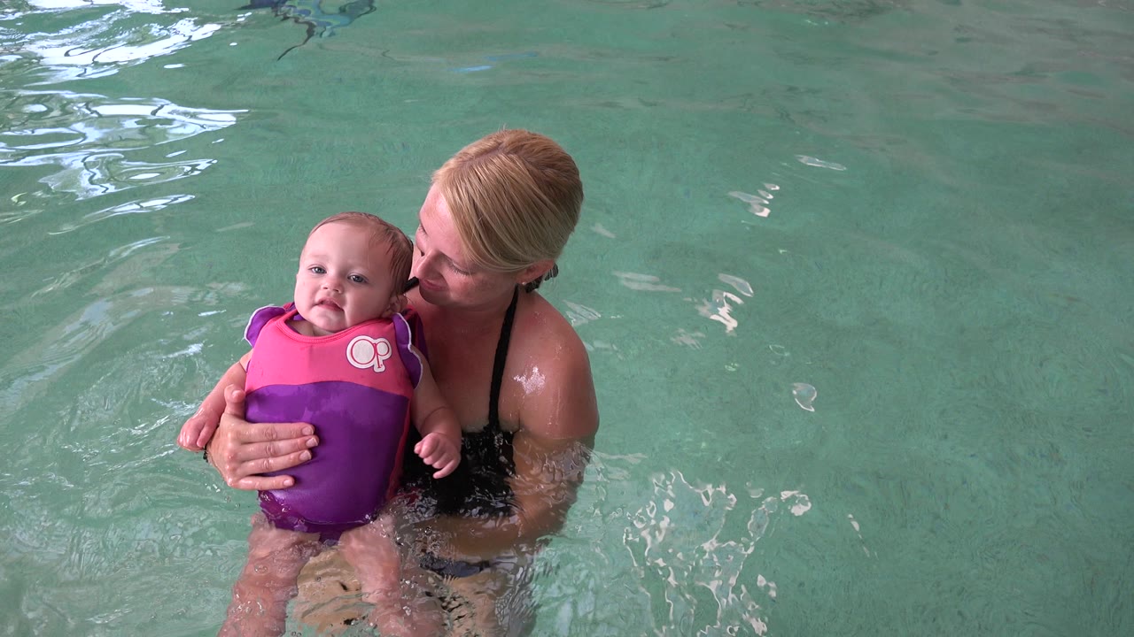 In Westminster, Colorado, the instructor or caregiver supports the infant from behind as the child faces forward in a bright flotation vest, surrounded by rippled pool water.