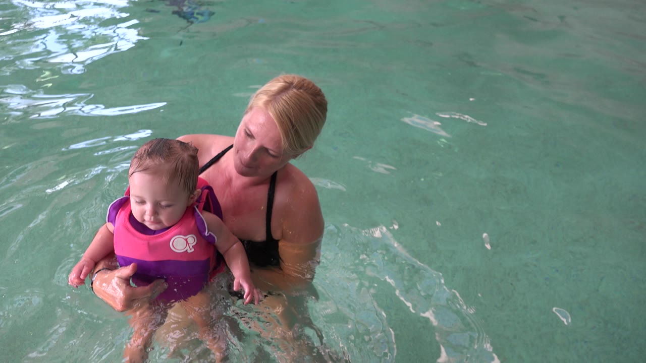 Infant in life vest looks down while woman steadies them in the pool.