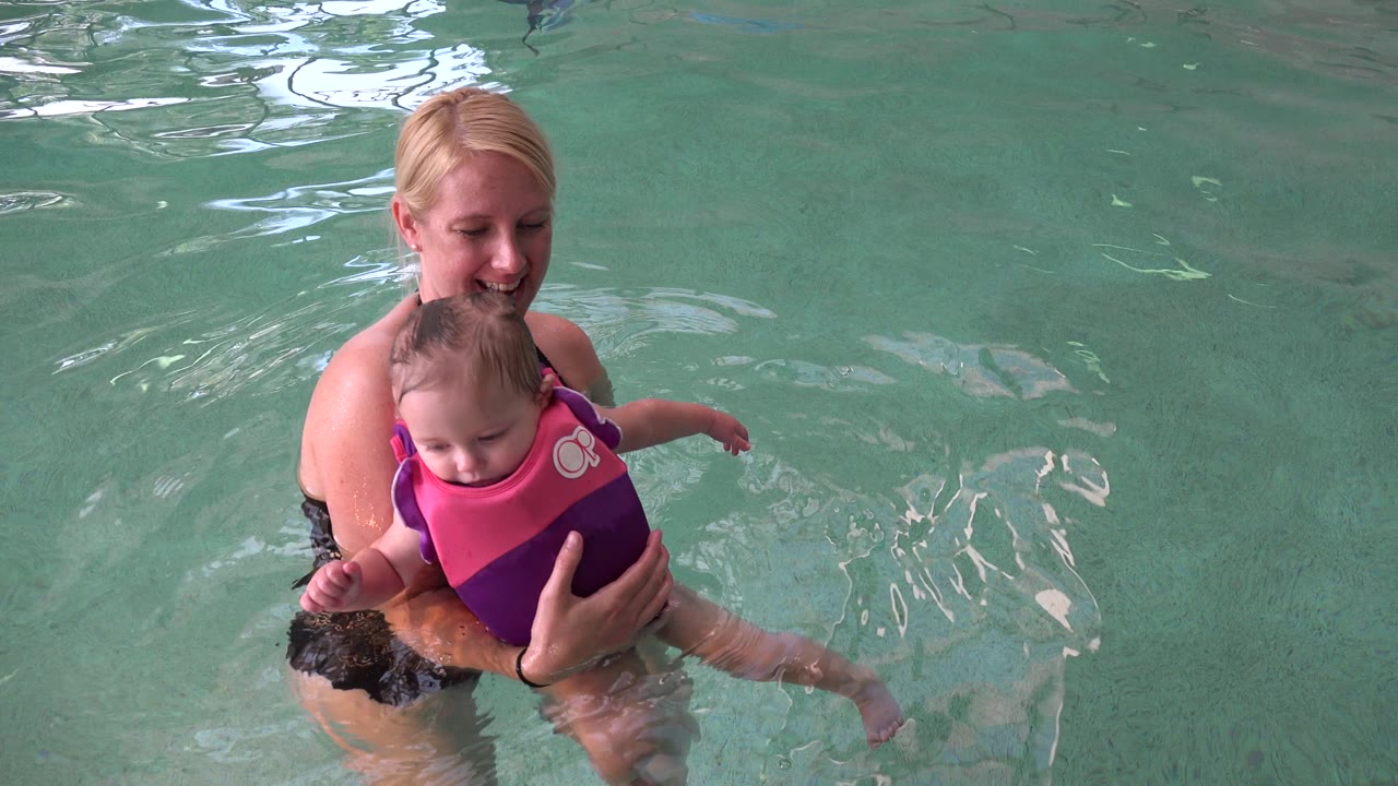 In Westminster, Colorado, a blonde woman in a black swimsuit supports a young child in a pink and purple life vest near the left side of the pool frame.