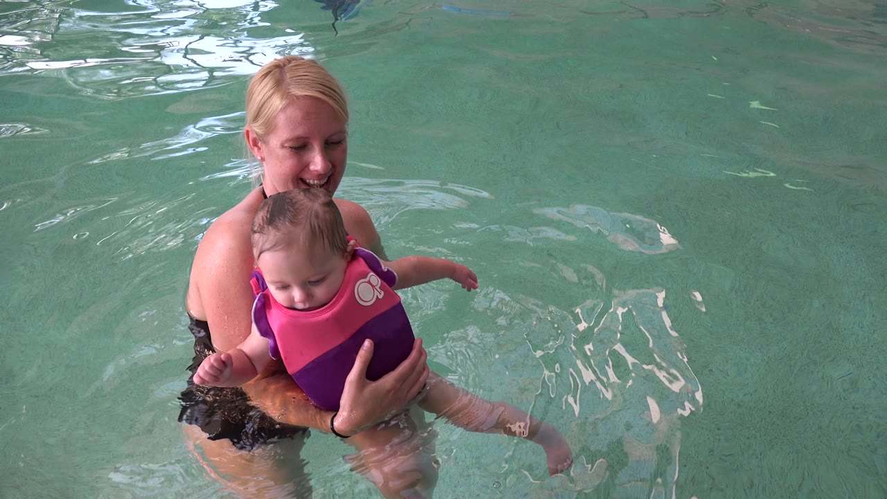 In Westminster, Colorado, the woman keeps both hands around the child as the child faces forward in a bright flotation vest and ripples spread across the pool surface.