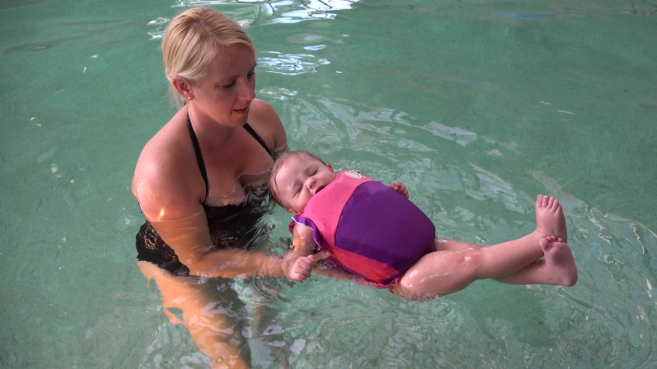The mother keeps one hand near the baby's head and torso while the infant floats face-up in a bright pink and purple vest in the Westminster pool.